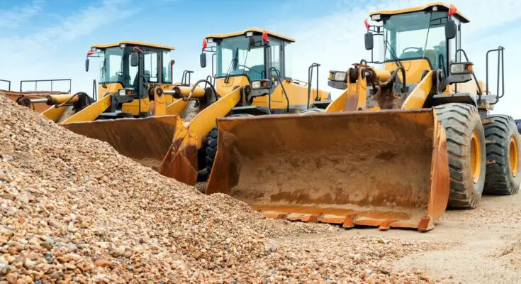 A row of three yellow excavators parked next to each other under a bright blue sky, and in front is a pile of rocks.
