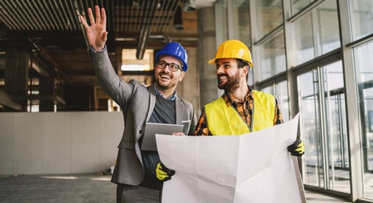 A man in a suit and hard hat gestures while standing beside a worker in a safety vest holding large architectural plans.