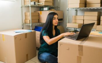 A young woman sits on the floor typing on a laptop that sits on a large cardboard box. Other boxes surround her.