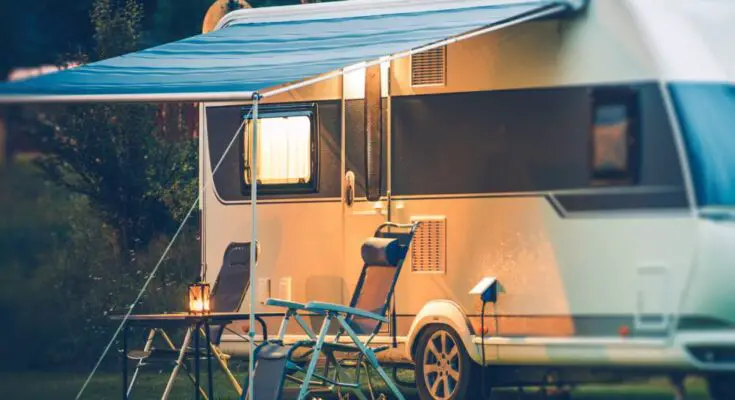 A small camper with a blue awning is parked on grass next to a pair of chairs and a small table with a lantern on it.