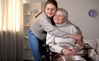 A young woman hugs an elderly woman in a wheelchair. Both women are smiling and have their eyes shut.