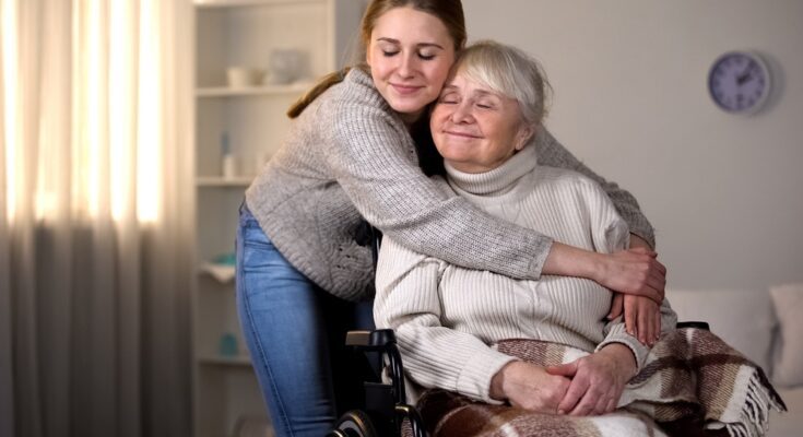 A young woman hugs an elderly woman in a wheelchair. Both women are smiling and have their eyes shut.