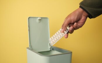 Someone holding medication who's about to dispose of it in a green trash bin. The wall in the background is yellow.