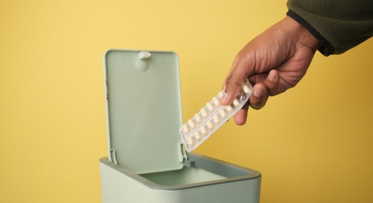 Someone holding medication who's about to dispose of it in a green trash bin. The wall in the background is yellow.