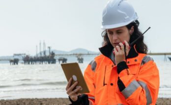 A woman in a high-visibility jacket and white hard hat looks at a tablet and speaks into a walkie-talkie.