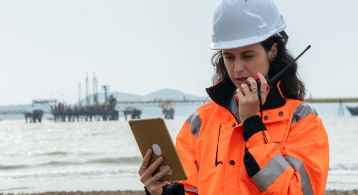 A woman in a high-visibility jacket and white hard hat looks at a tablet and speaks into a walkie-talkie.