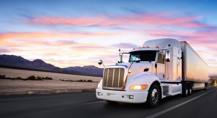 A white truck drives on a road. There's a desert, mountains, and a sunset surrounding the white truck.