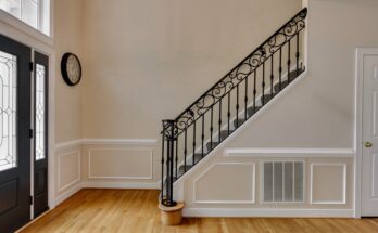 A home’s brightly lit front entryway features a staircase with an ornate metal banister. The entryway also has a glass door and a clock.