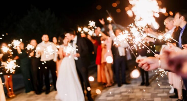 At a wedding, the bride and groom kiss while all of the other people around them hold out bright sparklers.
