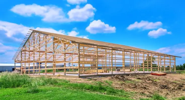 The wooden frame of a new pole barn is constructed on a cement foundation in a field on a sunny day with few clouds.