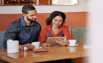 Two people in aprons sit at a table with coffee cups. They are both looking at a tablet screen.