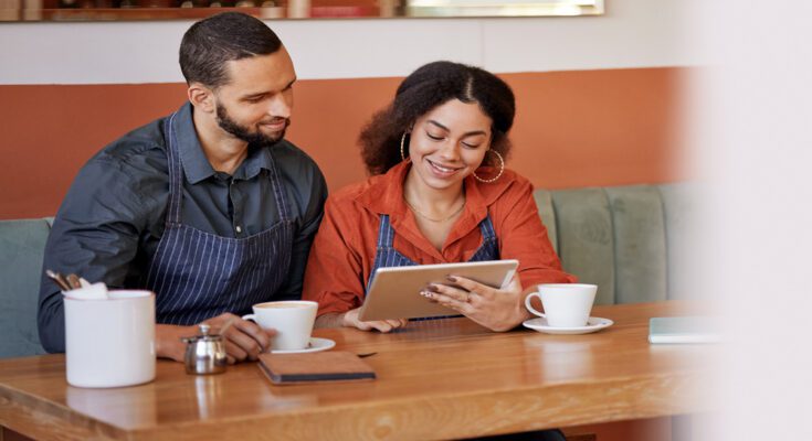 Two people in aprons sit at a table with coffee cups. They are both looking at a tablet screen.