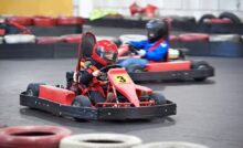 Two children are driving go-karts around an indoor track with bumpers. They're both wearing helmets.