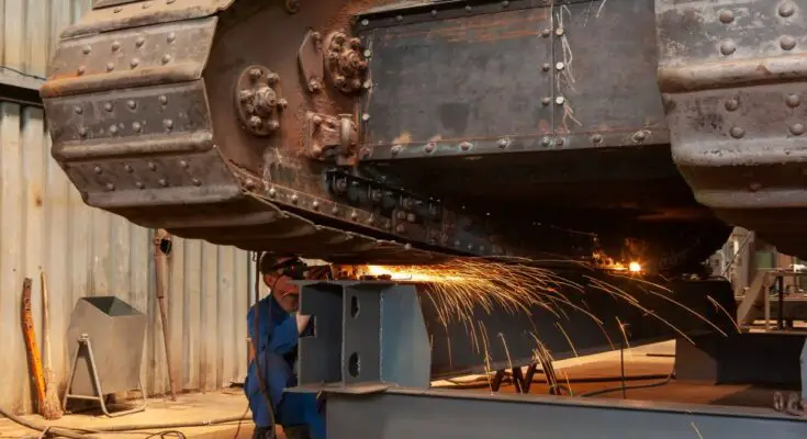 An industrial worker grinds on an old armored tank in a factory workshop. Sparks are flying off the metal.