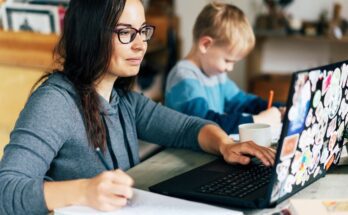 A woman works on a laptop and writes in a notebook as a young boy beside her draws with an orange pencil.