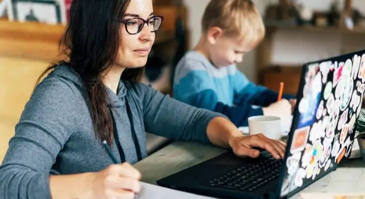A woman works on a laptop and writes in a notebook as a young boy beside her draws with an orange pencil.