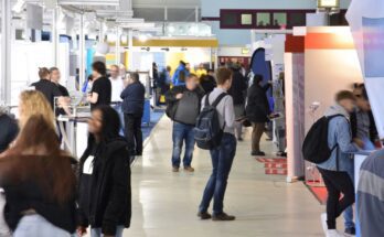 Countless, slightly blurred people walk around a trade show; some people stand in front of exhibit booths.