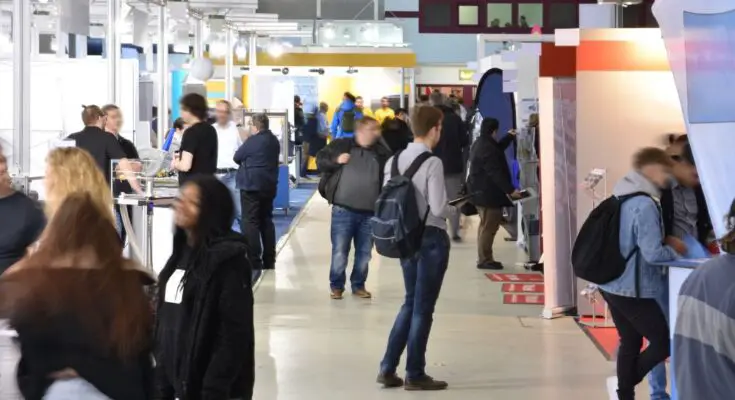 Countless, slightly blurred people walk around a trade show; some people stand in front of exhibit booths.