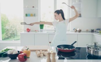 A woman stands in a martial arts-style pose as she cooks vegetables in her kitchen. She holds a spoon and a spatula.