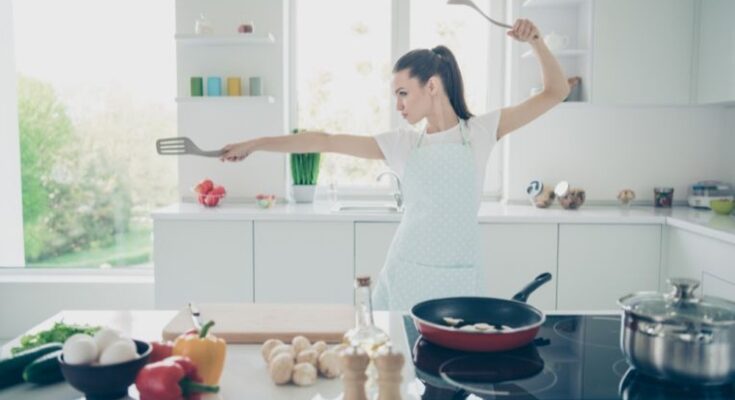A woman stands in a martial arts-style pose as she cooks vegetables in her kitchen. She holds a spoon and a spatula.