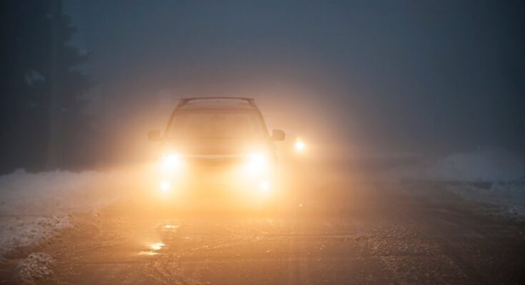 A close-up of the headlights of a car driving in fog. There's a bit of snow piled up on the side of the road.