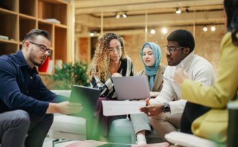 Three women and two men sit together looking over tablets and paperwork during a business meeting.