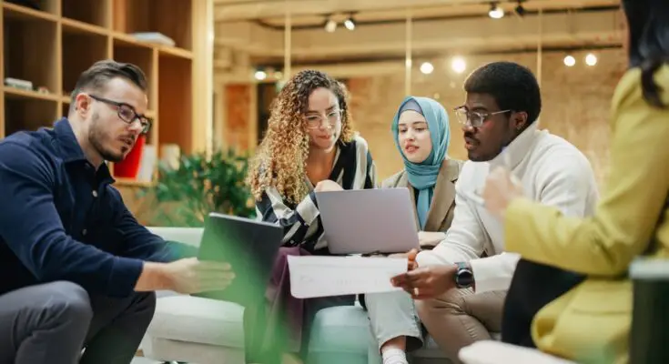 Three women and two men sit together looking over tablets and paperwork during a business meeting.