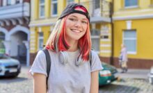 A teenage girl standing outside in an urban city. She has colorful dyed hair and headphones wrapped around her neck.