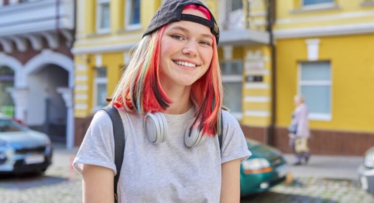 A teenage girl standing outside in an urban city. She has colorful dyed hair and headphones wrapped around her neck.