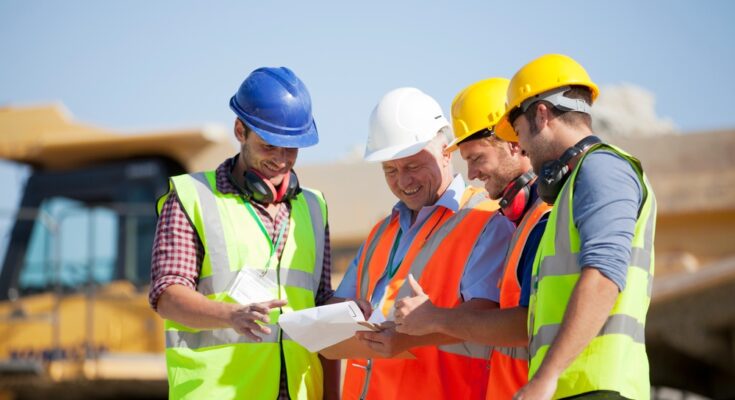 Four smiling construction crew members look at a clipboard together while standing on a job site on a sunny day.