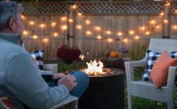 A man sitting in an outdoor chair next to a fire pit with string lights draped on a wooden fence in the background.