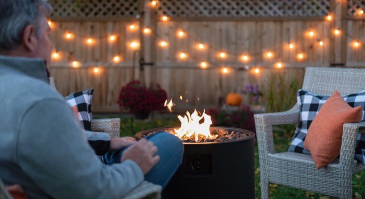 A man sitting in an outdoor chair next to a fire pit with string lights draped on a wooden fence in the background.