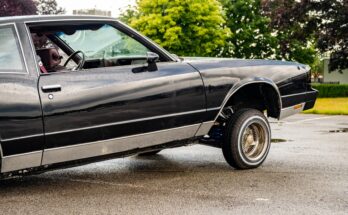 A glossy black vintage lowrider Cadillac-style car lifted up on wire wheels in an empty parking lot.