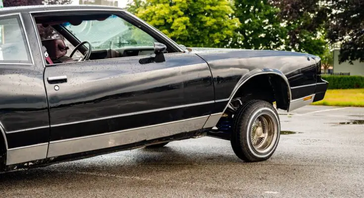 A glossy black vintage lowrider Cadillac-style car lifted up on wire wheels in an empty parking lot.