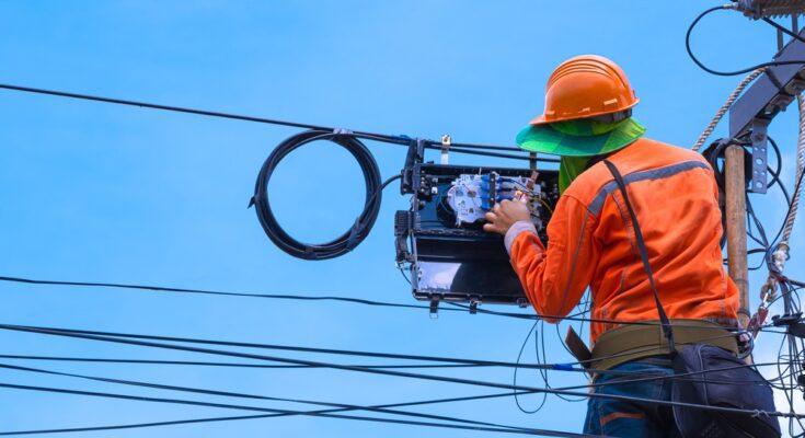 A technician checking over part of a utility line. He is wearing safety gear, a hard hat, and high-visibility clothes.