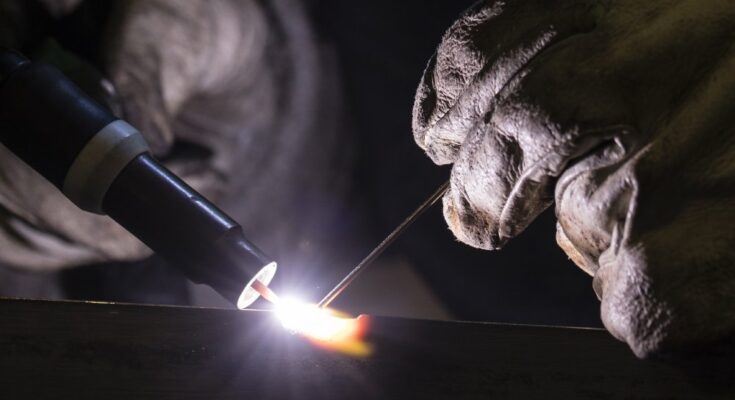 An extreme close-up shot of gloved hands using TIG (tungsten inert gas) welding, emitting a bright white light.