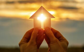 A pair of hands hold a piece of paper cut into the shape of a house with a cross in the middle. The sun shines through the cross.