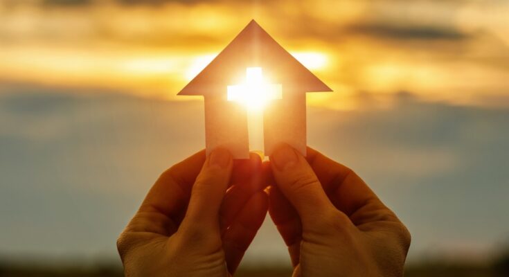 A pair of hands hold a piece of paper cut into the shape of a house with a cross in the middle. The sun shines through the cross.
