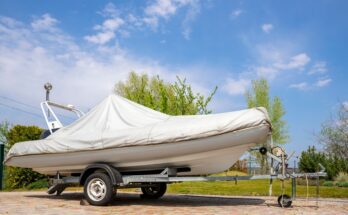 A boat with a white cover rests on a wheeled trailer on a sunny day, surrounded by trees in the background.