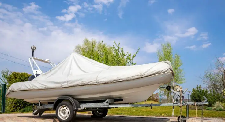 A boat with a white cover rests on a wheeled trailer on a sunny day, surrounded by trees in the background.