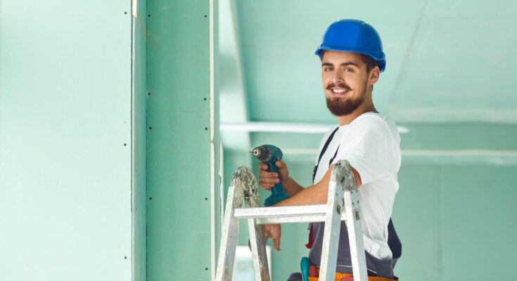 A man with a beard and a blue safety helmet, standing on a ladder, holding a drill to secure the sheets of drywall.