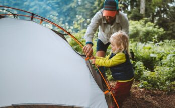 A man wearing a blue and orange hat crouches next to a child as they build a white tent outdoors.