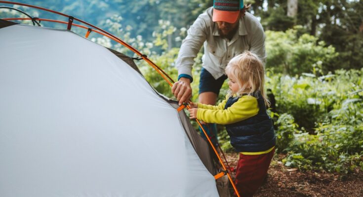 A man wearing a blue and orange hat crouches next to a child as they build a white tent outdoors.