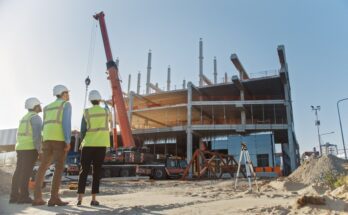Three workers in yellow safety vests and hard hats stand on a project site as workers and machines construct a building.