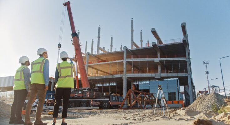 Three workers in yellow safety vests and hard hats stand on a project site as workers and machines construct a building.