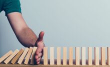 A person holding their hand in the way of a series of falling wooden blocks to protect other standing blocks.