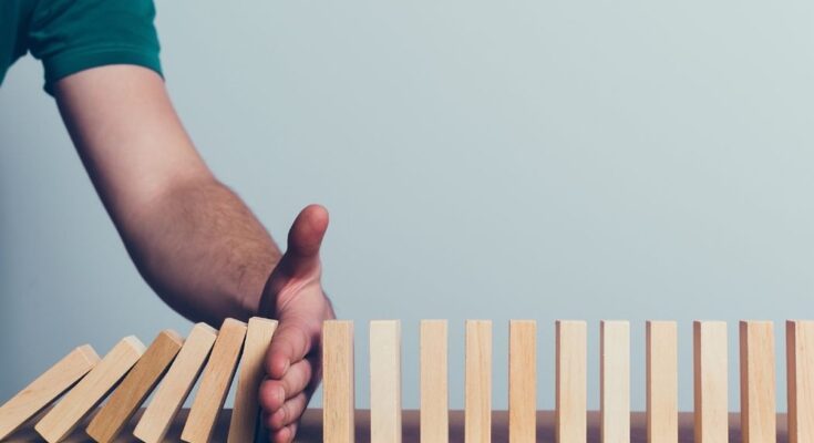 A person holding their hand in the way of a series of falling wooden blocks to protect other standing blocks.