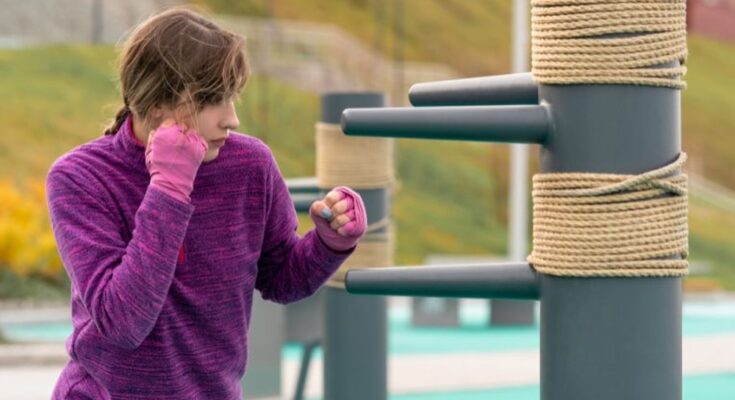 A woman practicing martial arts with a traditional dummy. She is wearing pink bindings on her hands and a purple sweater.