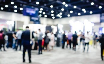 A blurred view of a large conference event with people walking and standing around. The lights are bright above.