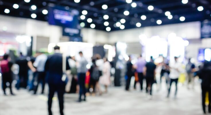 A blurred view of a large conference event with people walking and standing around. The lights are bright above.